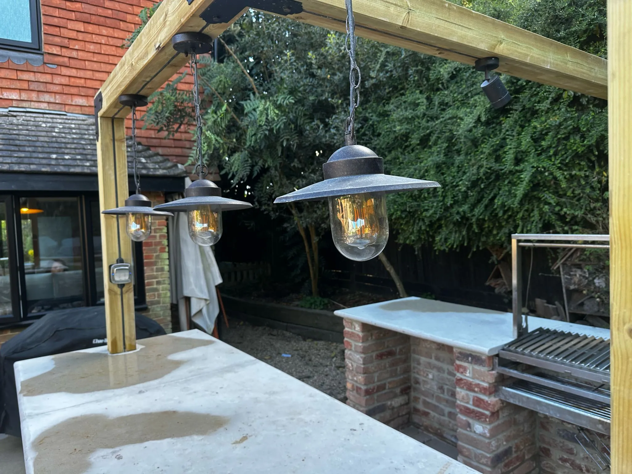 Three garden lights, hung above an outdoor kitchen worktop, with more worktop space and a grill also visible